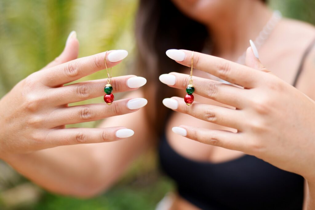 Woman showing off white almond nails while holding red and green earrings — a stylish example of the almond nail shape trend.