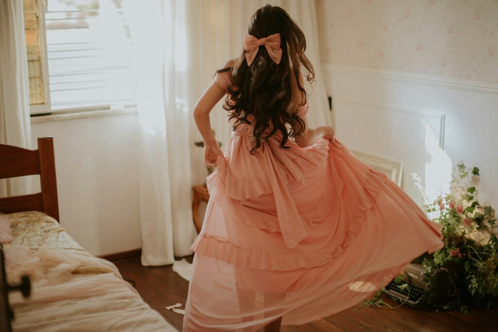Woman in a soft pink dress representing the playful and stylish appeal of shein dresses, standing by a bed in a modern, minimalist room