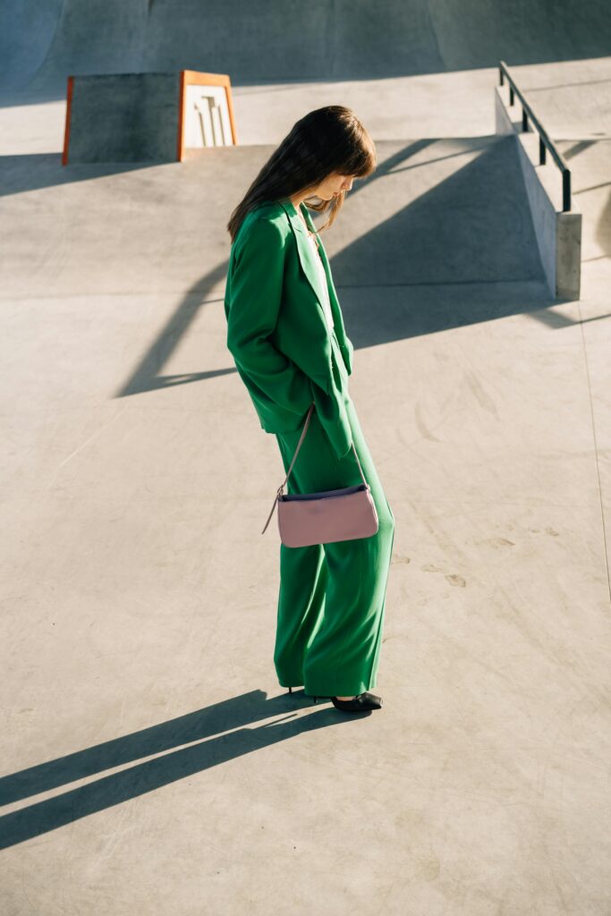 Woman in a green suit carrying a pink saddle bag while walking outdoors, showcasing a modern and stylish accessory look.