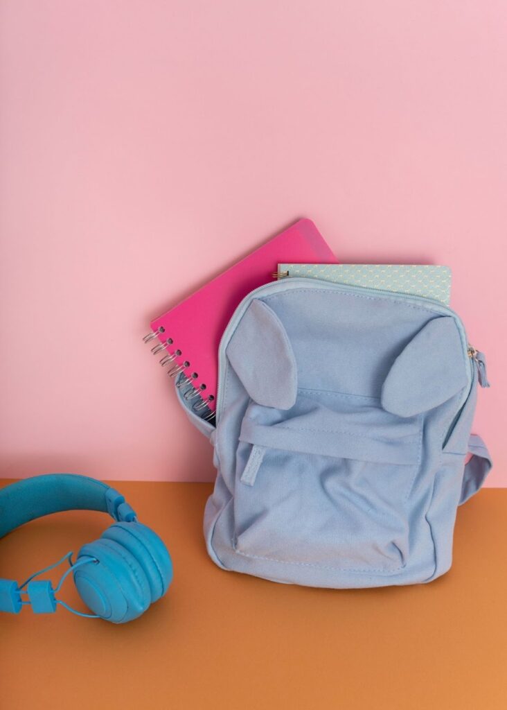 Preppy backpacks styled with colorful notebooks and headphones on a pink and orange background.