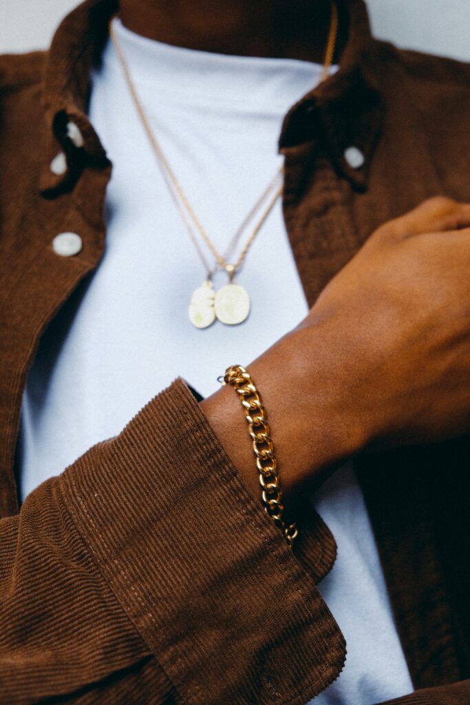Close-up of a man wearing a chunky gold bracelet and layered gold necklaces with a casual brown jacket and white shirt.
