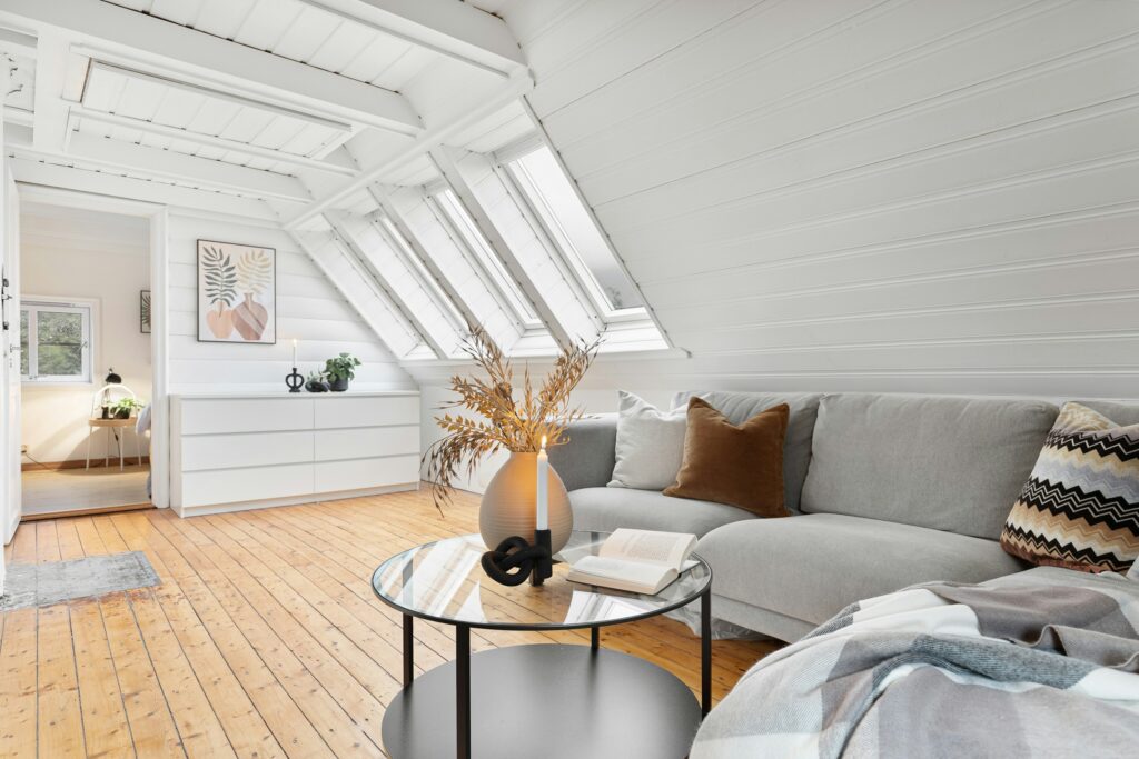 Cozy modern living room with dramatic white vaulted ceiling, exposed beams, and multiple skylights flooding the space with natural light. A plush gray sectional sofa, warm wood floors, and stylish neutral decor create an airy yet inviting Scandinavian-inspired retreat.