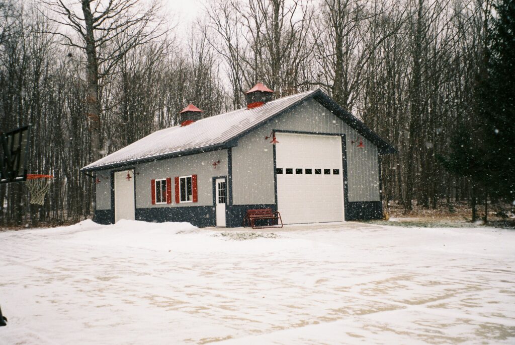 Snowy detached garage with a white insulated-style garage door and small upper windows on a winter day with light snow falling – perfect example of a home that benefits from garage door insulation in cold climates.