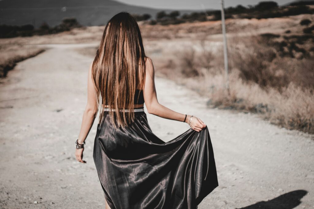 A woman with long, straight brown hair walks away from the camera on a dusty rural road at golden hour. She wears a long black satin slip dress that catches the light beautifully, holding the flowing hem in one hand to lift it slightly as she moves.
