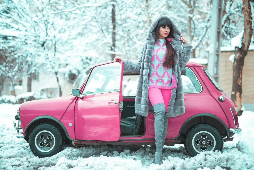 Woman in vibrant pink winter outfit female leaning on a pink car in the snow, featuring a faux fur coat over a pink heart-patterned sweater and pink leggings.