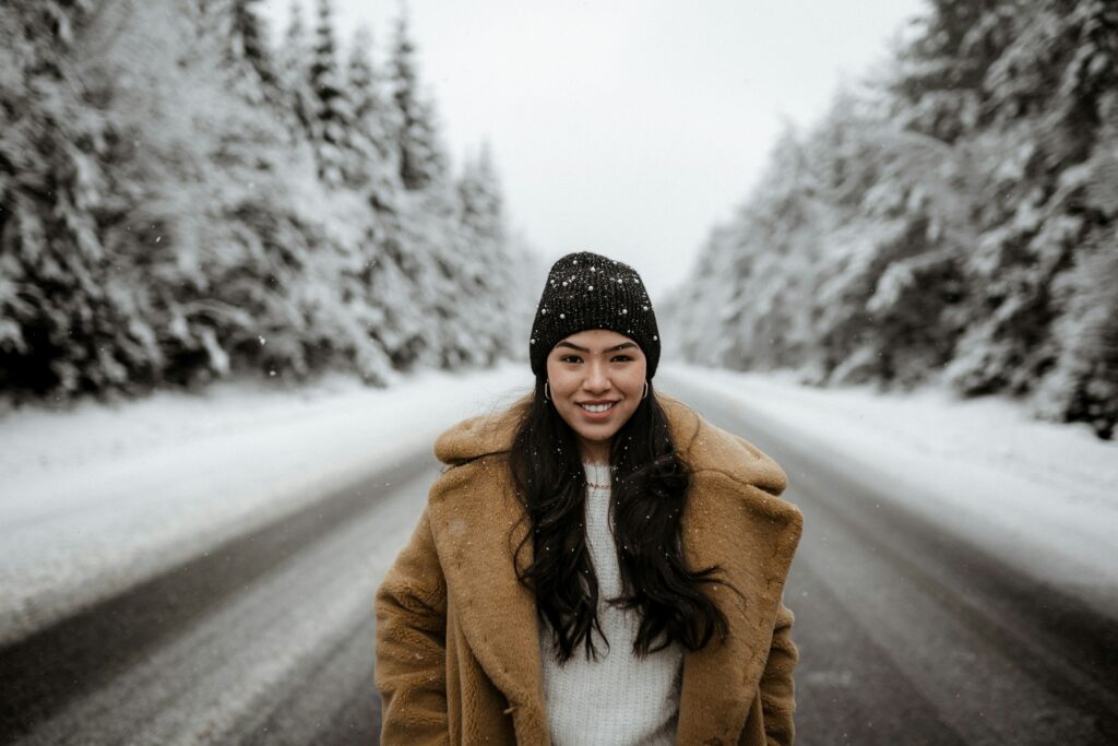 Young woman in a trendy winter outfit female: camel teddy coat over white sweater, black beanie with pearls, smiling in falling snow on a snowy forest road.