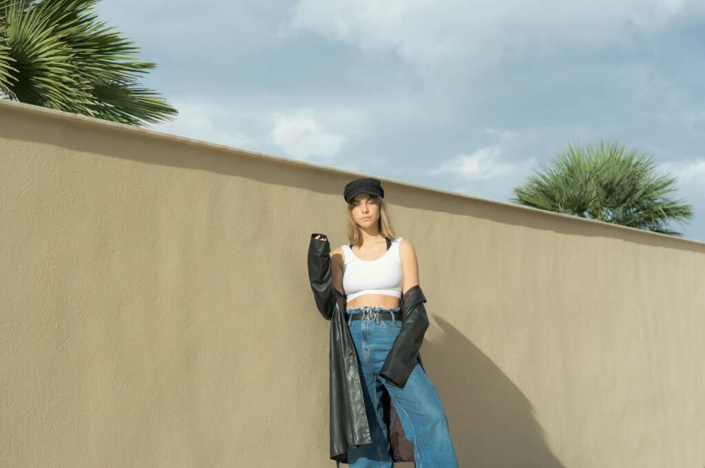 Young woman wearing fitted white crop top, wide-leg baggy blue jeans, oversized black leather jacket draped loosely, black cap, standing in sunny outdoor setting with palm trees and beige wall.