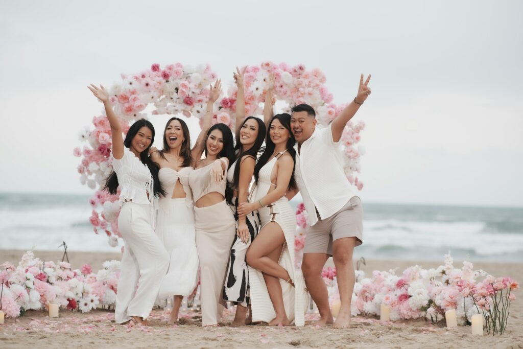 Smiling friends posing at a chic beach bachelorette party with pink rose floral arch and scattered petals.