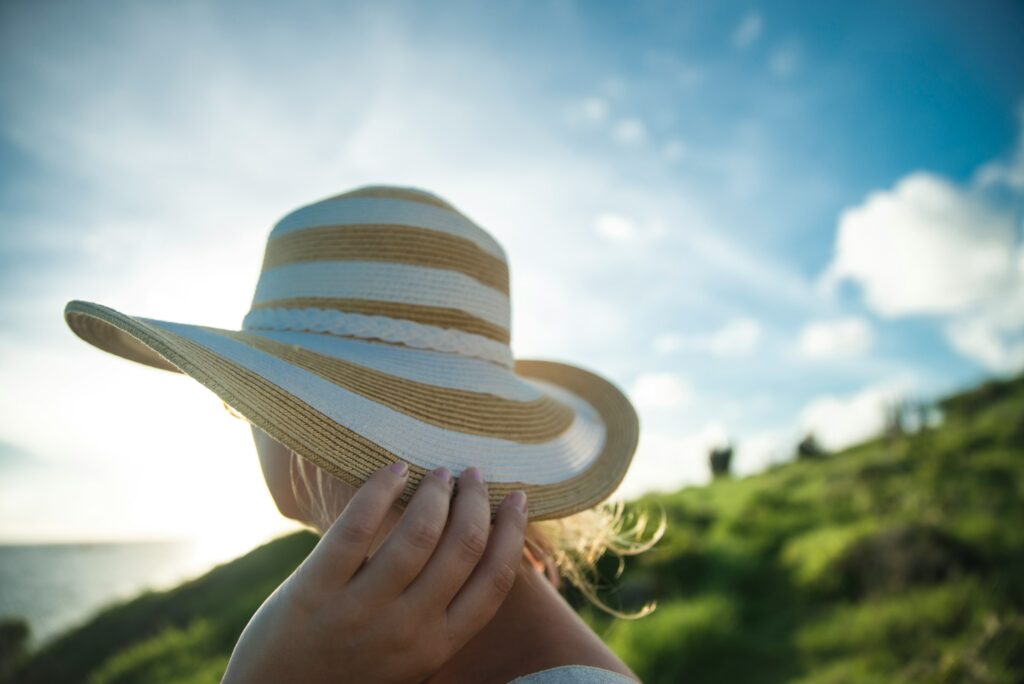 A woman wearing a yellow and white striped wide-brim hat, holding the brim while enjoying a sunny coastal view with blue sky and ocean in the background, capturing the effortless summer 2026 style of the striped hat trend.