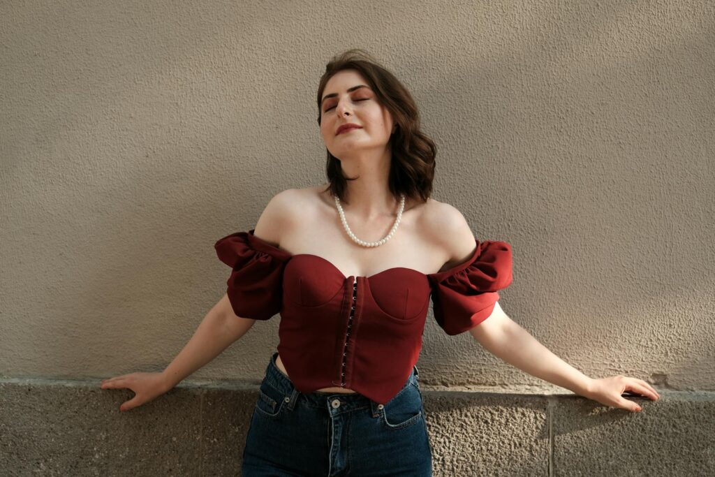 Woman wearing a burgundy sweetheart neckline top with puff sleeves styled with blue jeans against a neutral wall.
