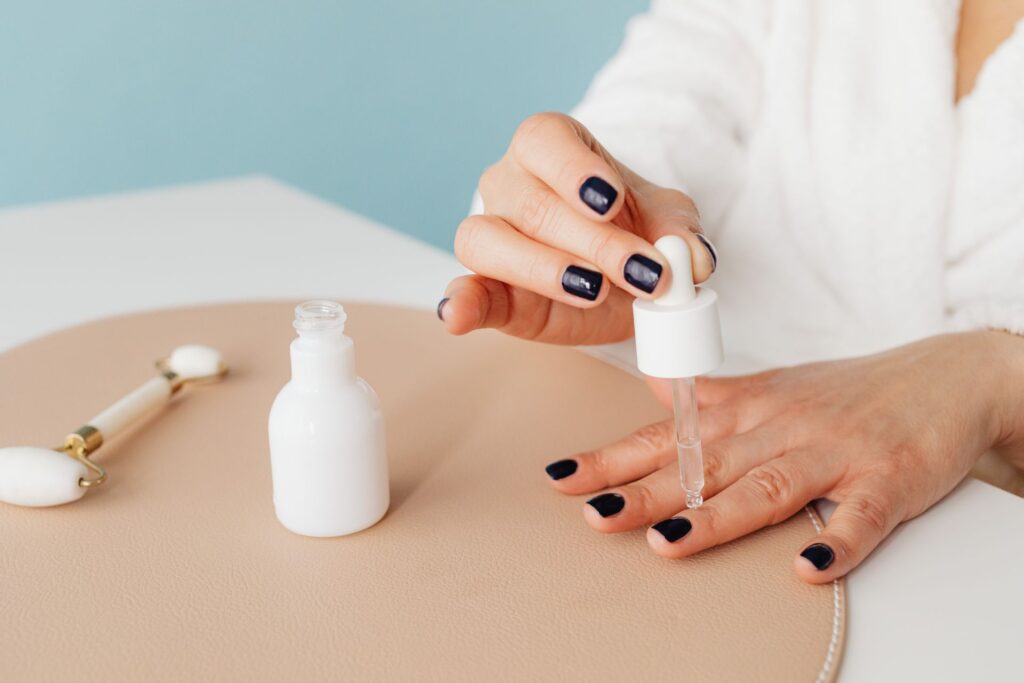 Woman applying cuticle remover serum with dropper on dark nails at vanity.