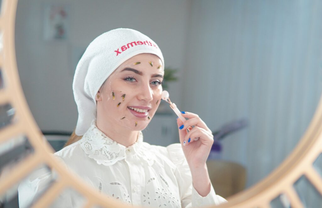 A woman using a rose quartz facial roller at home during a Korean skin treatment with botanical face mask and hair wrapped in a white towel
