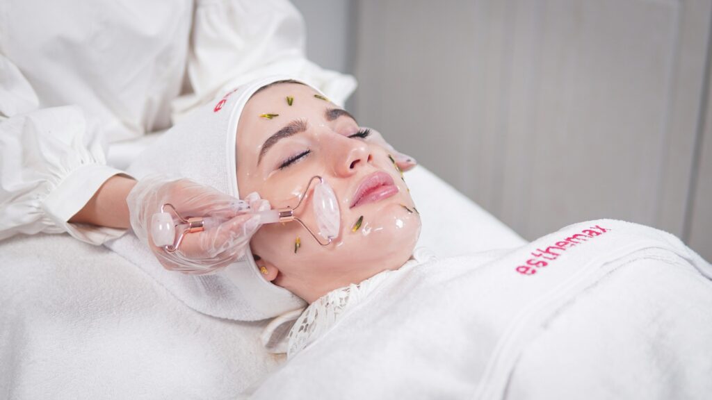 A woman receiving a professional Korean skin treatment at a beauty clinic with a rose quartz facial roller, hydrating mask, and botanical ingredients applied to the face.