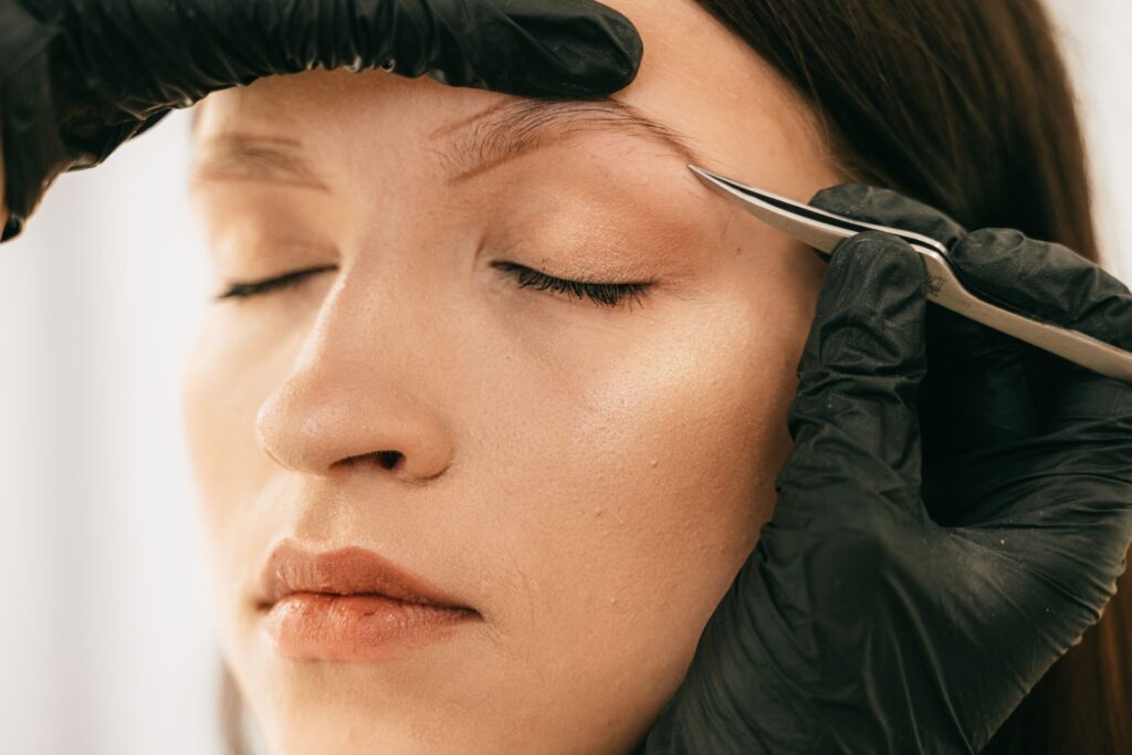 Professional esthetician using tweezers to shape eyebrows on a woman with gloved hands.