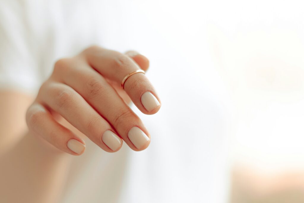 Woman with short gel manicure in nude beige on rounded nails with gold ring.