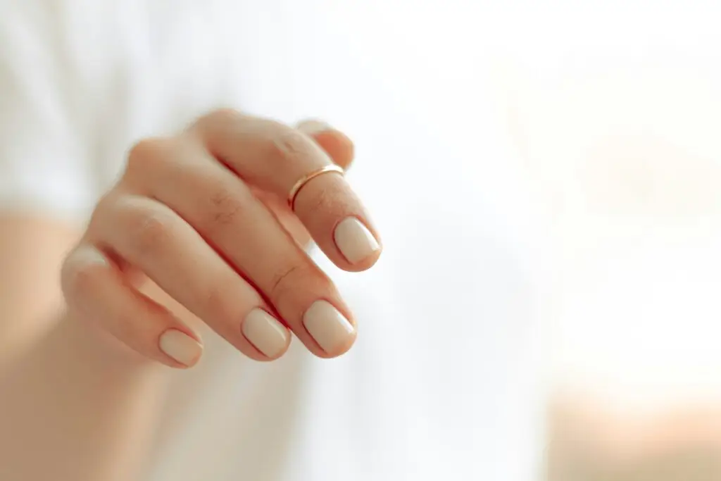 Woman with short gel manicure in nude beige on rounded nails with gold ring.