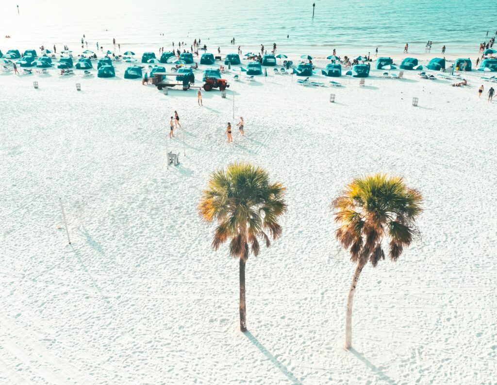 Aerial view of a crowded Gulf Coast beach in Florida with palm trees, beach cabanas, and people playing volleyball. 
