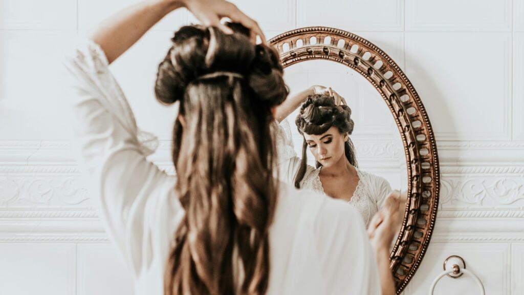Woman styling an elevated twisted updo with loose curls in a mirror.