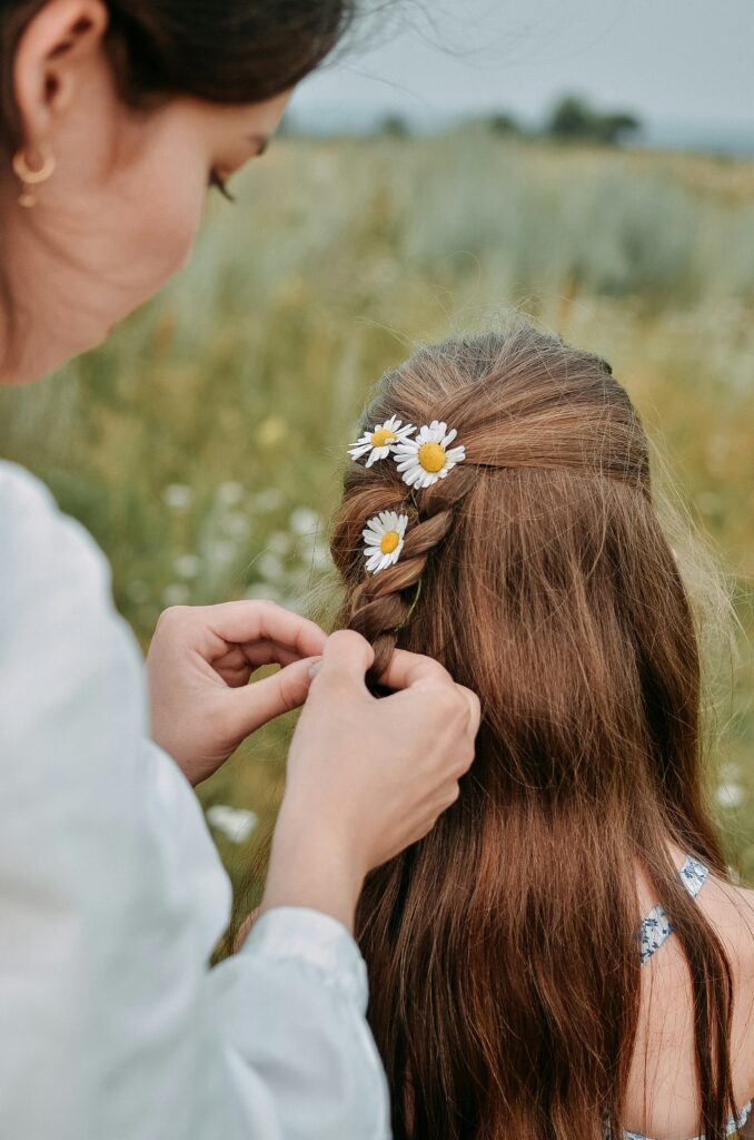 Woman creating a floral boho braid hairstyle with fresh daisy flowers woven into long brown hair.