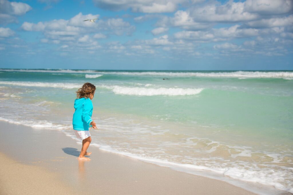 Toddler in a turquoise rash guard wading in shallow, calm waves on a Florida beach.