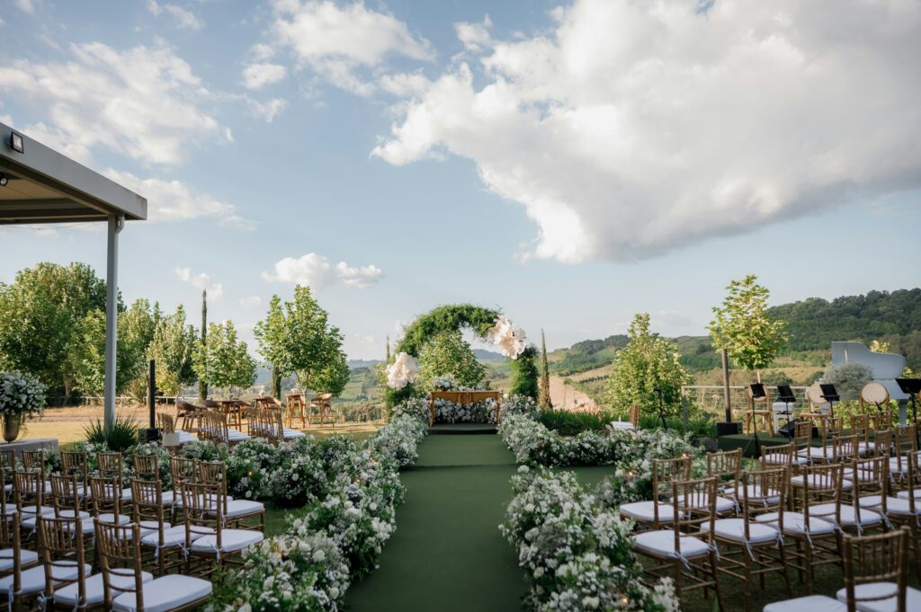 Outdoor wedding ceremony aisle with white floral arch, wooden chairs, and rolling hillside view.