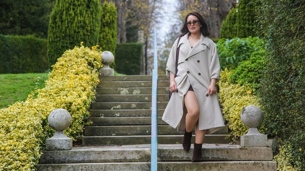 Woman wearing a beige double-breasted trench coat over a short dress with brown ankle boots on garden steps.