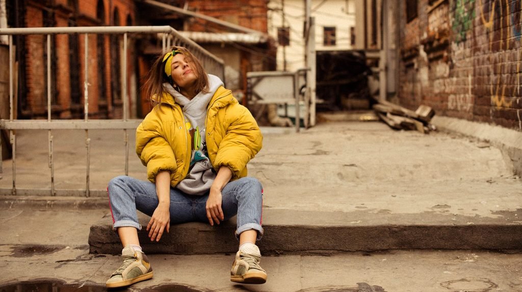 Woman in a mustard-yellow puffer jacket over a gray hoodie and light-wash jeans, sitting on a concrete step in a brick alley.